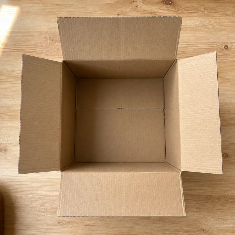 An empty open cardboard box seen from above on a wooden floor.